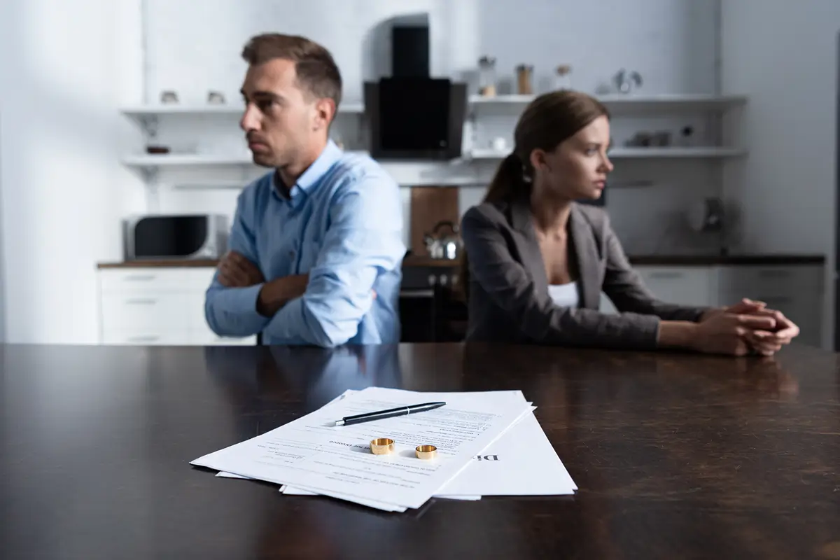 Couple sitting apart at a table with divorce papers and wedding rings in focus, symbolizing separation.