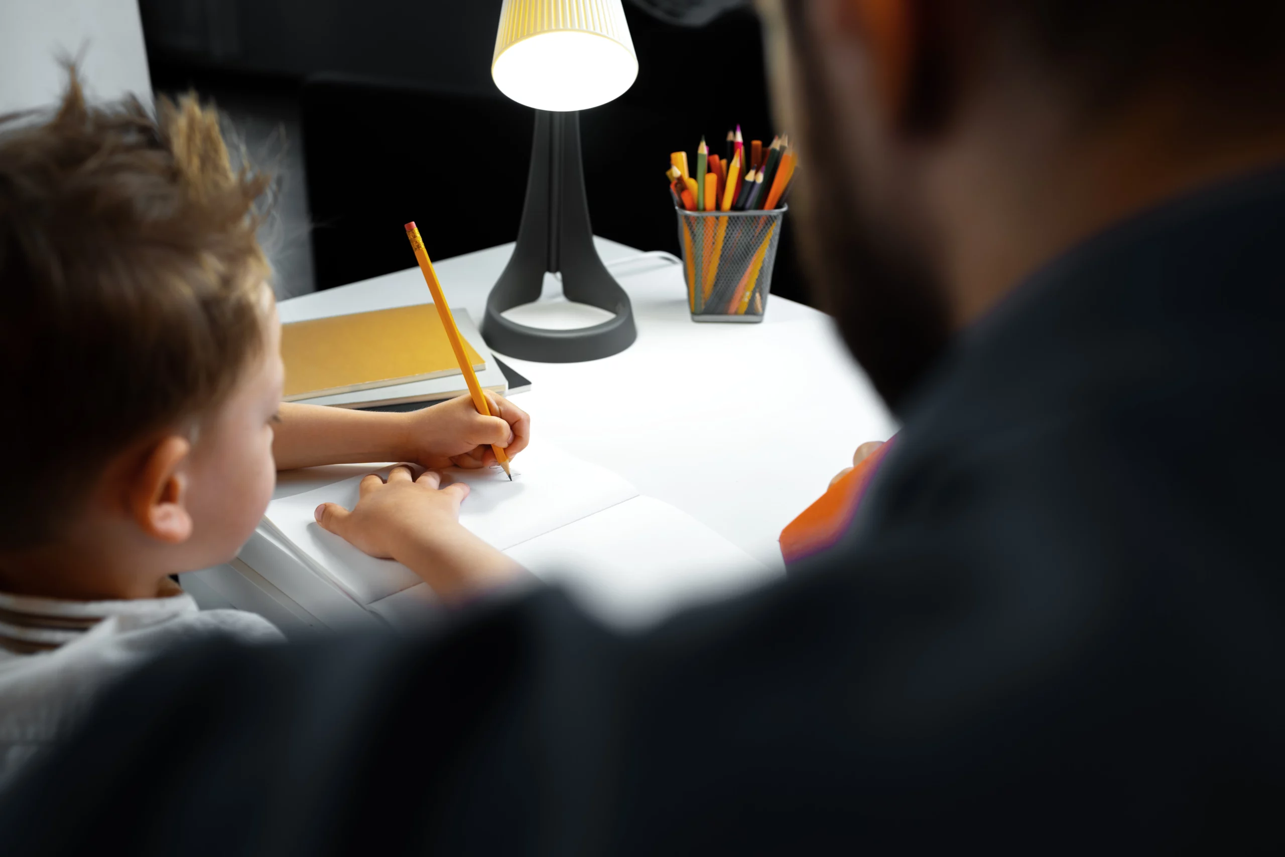 A child guided by an adult writes in a notebook at a well-lit desk, surrounded by books and colored pencils — symbolizing the caregiving and legal guardianship process in California.