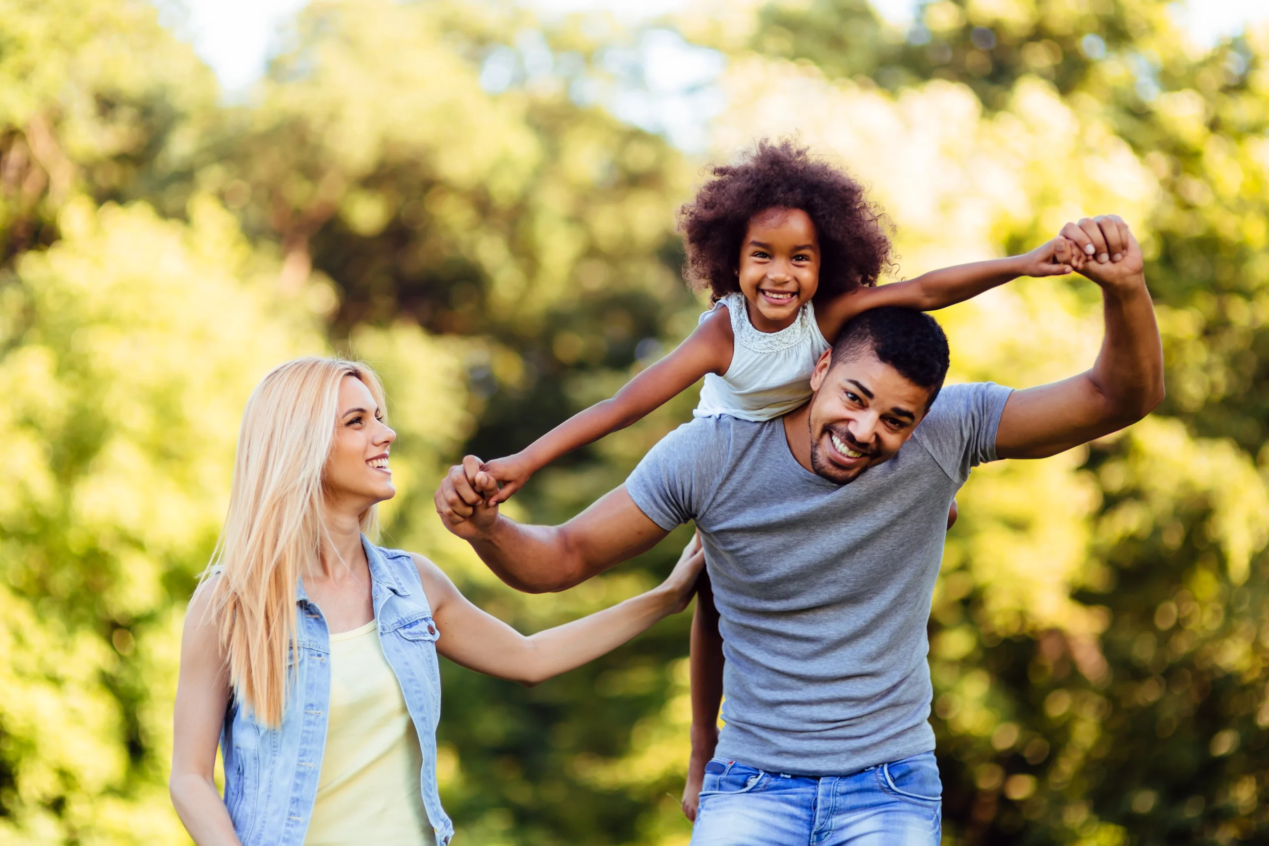 Joyful family in a sunny park: child on father’s shoulders holding mother’s hands, greenery in the background.